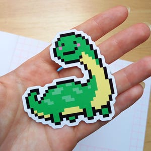 Könnte beinhalten: Ein grüner Pixel-Art-Dinosaurier-Aufkleber mit gelbem Hals und lächelndem Gesicht. Der Aufkleber hat eine schwarze Umrandung und befindet sich auf weißem Hintergrund. Der Dinosaurier posiert.