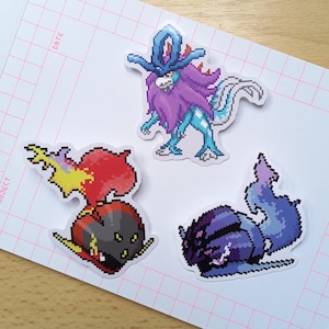 Könnte beinhalten: Drei Pixel-Art-Aufkleber mit legendären Kreaturen. Einer zeigt ein lila-blaues Wesen mit langem Schwanz und Krone. Ein anderer zeigt ein rot-schwarzes Wesen mit Flammen. Der dritte Aufkleber zeigt ein lila-blaues Wesen.