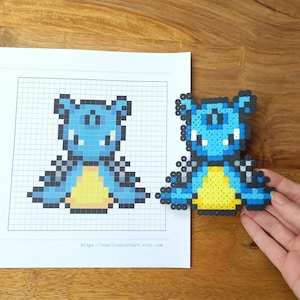 K&ouml;nnte beinhalten: Eine Pixel-Art-Figur aus blauen, gelben und schwarzen Kunststoffperlen, neben einem auf Papier gedruckten Rastermuster. Das Papier enth&auml;lt den Text https://snailventureart.etsy.com und ein passendes Rasterdesign der gleichen Figur.