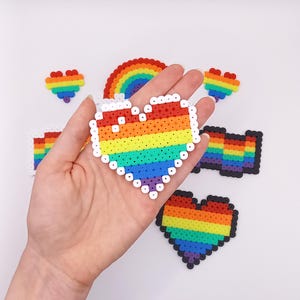 Könnte beinhalten: Ein Regenbogenherz, Regenbogenstreifen und ein Regenbogenherz mit schwarzem Umriss, alles aus bunten Perlen.