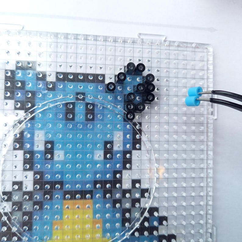 K&ouml;nnte beinhalten: Eine durchsichtige, quadratische Steckplatte enth&auml;lt ein teilweise zusammengesetztes Pixel-Art-Design aus kleinen Kunststoffperlen. Das Design zeigt Blau-, Schwarz- und Gelbt&ouml;ne. Eine schwarze Pinzette h&auml;lt zwei hellblaue Perlen am rechten Rand.