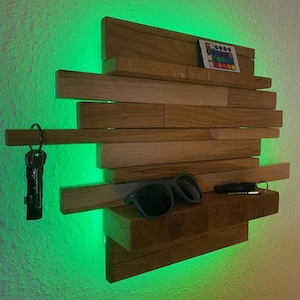 Può includere: Mensola da parete in legno con un design a strati, che contiene occhiali da sole, chiavi e un telecomando. La mensola è illuminata da luci a LED verdi, creando un look moderno. Le chiavi sono attaccate a un portachiavi nero.