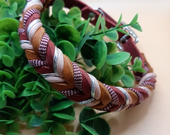May include: A braided leather dog collar with brown, pink, and silver stripes. The collar is lying on a bed of green leaves.