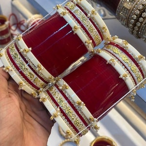 Può includere: Un set di braccialetti rossi e bianchi con dettagli dorati e piccole perle. I braccialetti presentano un design ornato con una finitura lucida. Altri braccialetti sono visibili sullo sfondo, in stile tradizionale.