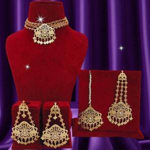 May include: A gold-tone jewelry set with a choker necklace, earrings, and a maang tikka. The necklace has a large, ornate pendant with a floral design. The earrings and maang tikka have similar designs with dangling chains and beads.