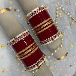 Può includere: Due braccialetti rossi e dorati con disegni intricati, esposti su un supporto cilindrico di colore neutro. I braccialetti presentano dettagli dorati e dettagli bianchi e dorati. Una collana di perle è drappeggiata vicino.