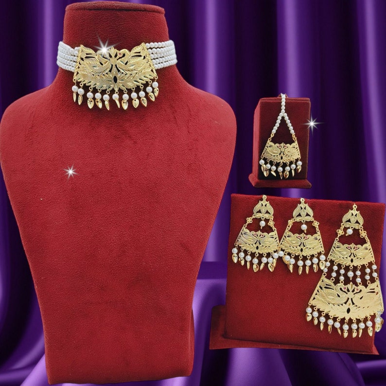 May include: A white pearl necklace with a gold pendant, a gold tikka, and a pair of gold earrings with pearl accents. The jewelry is displayed on red velvet.