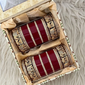 Puede incluir: Un conjunto de cuatro brazaletes rojos y dorados con diseños intrincados. Los brazaletes están en una caja con un patrón floral.