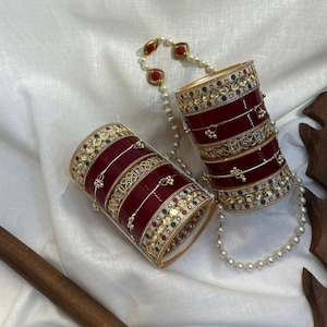 Può includere: Due set di braccialetti rossi e dorati ornati, con dettagli intricati e pietre preziose incastonate. Una collana di perle con accenti rossi collega i set. I braccialetti poggiano su un tessuto bianco.