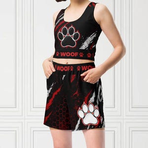 Op de afbeelding: Zwarte en rode atletische crop top en short set met een pootafdruk ontwerp. De top heeft een pootafdruk grafiek en het woord "WOOF" erop gedrukt. De shorts hebben ook een pootafdruk grafiek.