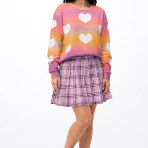 May include: A long-sleeve crewneck sweatshirt featuring a pink, orange, and yellow gradient background with white heart patterns, paired with a light purple pleated plaid skirt and black lace-up boots. The text MAX(XX)ART and SOLD SEPARATELY appear.