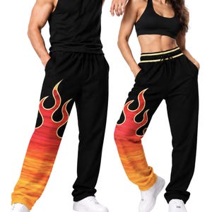 Puede incluir: Pantalones de chándal negros con un gráfico de llamas en rojo, naranja y amarillo en la parte inferior de la pierna izquierda. Los pantalones tienen una cintura elástica con cordón. El texto MAX(XX)ART es visible en la esquina superior izquierda.
