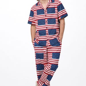 May include: A short-sleeved button-down shirt and matching pants set featuring a repeating pattern of the American flag with red and white horizontal stripes and blue rectangles with white stars. The set includes white slide sandals.