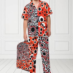 Puede incluir: Un conjunto de camisa y pantalón con estampado rojo y negro con círculos blancos. La camisa tiene cuello y mangas cortas. El pantalón es largo y tiene pierna ancha. Una maleta negra con el mismo estampado está en el fondo.
