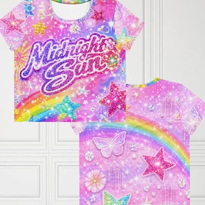 Op de afbeelding: Een crop top met korte mouwen met een levendig, kleurrijk ontwerp. De shirt heeft een regenboog, sterren, vlinders en de tekst "Midnight Sun" in een glitterlettertype. De achtergrond is een mix van roze, paars en blauw.