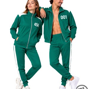 May include: A green track jacket and matching track pants set featuring white side stripes and a white zip. The jacket displays the number 456 or 001 in white on the chest. The set includes a high collar, ribbed cuffs, and a straight-leg fit.