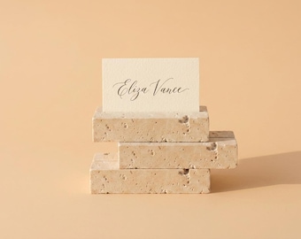 May include: A rectangular cream-colored card with the name Eliza Vance printed in black cursive script, held upright by a stack of three rectangular beige travertine stone blocks with a porous, textured surface and natural pitted details.