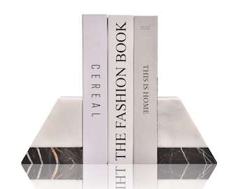 Könnte beinhalten: Drei Bücher mit den Titeln Cereal, The Fashion Book und This Is Home stehen zwischen zwei trapezförmigen Buchstützen. Die Buchstützen bestehen aus einem oberen Teil aus weißem Marmor und einem schwarzen Marmorsockel mit weißen und orangen Adern.