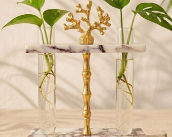 May include: A propagation station featuring two clear glass test tubes held between two white and purple marble slabs. A central gold-toned metal rod connects the slabs, topped with a gold-toned coral-shaped ornament. Green plants are inside.