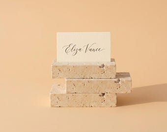 May include: A rectangular cream-colored card with the name Eliza Vance printed in black cursive script, held upright by a stack of three rectangular beige travertine stone blocks with a porous, textured surface and natural pitted details.