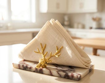May include: A rectangular white marble base with dark purple veining features a gold-toned metal deer head figurine attached to the front. A folded beige linen napkin is held upright by the deer antlers, creating a functional table accessory.