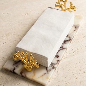 May include: A rectangular white marble tray with dark brown veining, featuring two gold-coloured metal handles shaped like coral branches. A stack of folded white paper napkins is positioned between the two decorative gold coral handles.