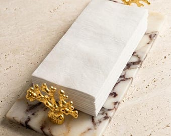 May include: A rectangular white marble tray with dark brown veining, featuring two gold-colored metal handles shaped like coral branches. A stack of folded white paper napkins is positioned between the two decorative gold coral handles.