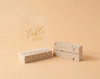 Könnte beinhalten: Ein durchsichtiges, rechteckiges Acrylschild mit dem goldenen Schriftzug "table ONE", das in einen beigen, porösen Steinsockel eingesetzt ist, begleitet von einem zweiten identischen Steinsockel mit einem horizontalen Schlitz.