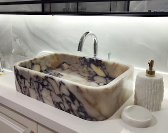 Könnte beinhalten: Ein rechteckiges Aufsatzwaschbecken aus Marmor mit weißer und dunkelvioletter Maserung sowie einem Chrom-Wasserhahn. Daneben stehen ein weißer, strukturierter Seifenspender mit goldener Pumpe und eine runde, weiße Seifenschale auf einer Ablage.