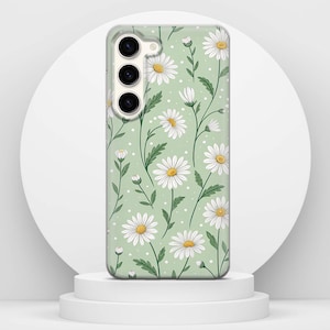 May include: A phone case with a daisy floral design. The case has a light green background with white daisies and yellow centres. Green stems and leaves are also present. The phone case is on a white pedestal.