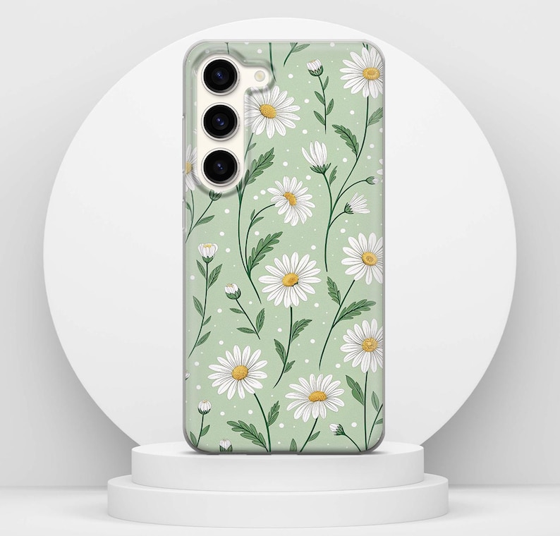 May include: A phone case with a daisy floral design. The case has a light green background with white daisies and yellow centres. Green stems and leaves are also present. The phone case is on a white pedestal.