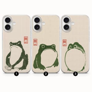 May include: Three phone cases with a cream background and green frog illustrations. Each case features a different frog design, with one holding a bowl. The cases also have a red Japanese character in the upper left corner.