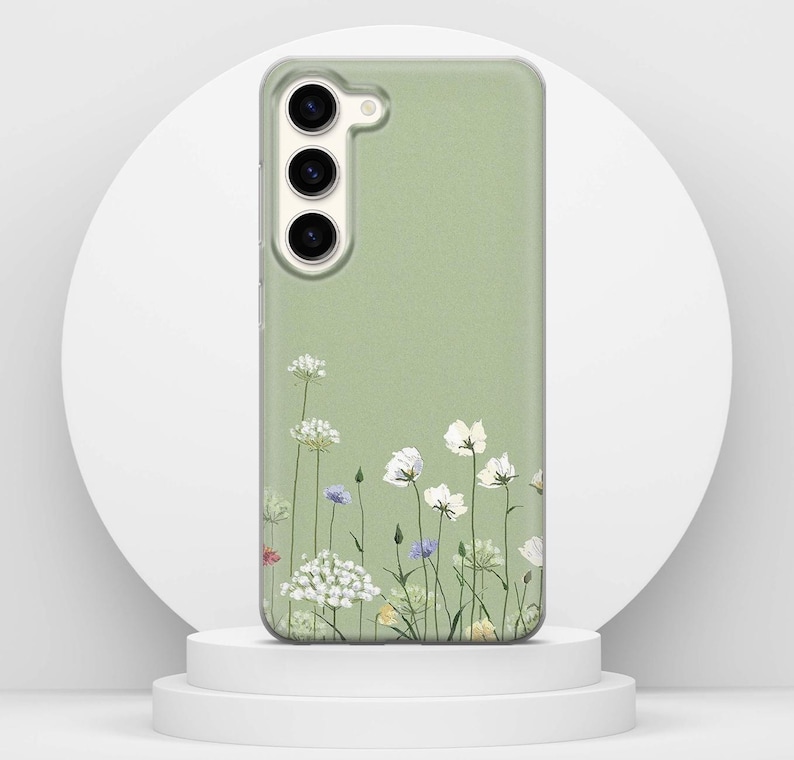 May include: A sage green phone case with a floral pattern. White, blue, and yellow wildflowers are shown at the base of the case. The phone case has a camera cutout and buttons.
