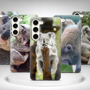 May include: A collection of phone cases featuring koala bear designs. The cases display close-up images of koalas in various poses, including one clinging to a tree trunk. The cases have a glossy finish and are displayed on a white platform.