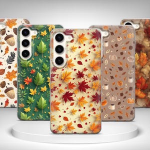 Puede incluir: Una colección de fundas para teléfonos con diseños otoñales. Las fundas presentan patrones de hojas, bellotas y calabazas en tonos rojos, naranjas, verdes y marrones. Las fundas se exhiben sobre una plataforma blanca y circular.
