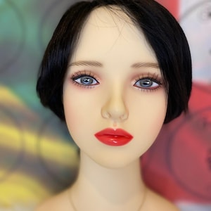 Puede incluir: Un maniquí con el pelo negro corto, que lleva un collar de oro con la palabra "PRISTINE" escrita. El maniquí tiene lápiz labial rojo y ojos azules.