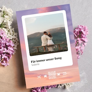 Könnte beinhalten: Eine Karte mit einem Foto eines Paares, das sich auf einer Bank vor einer Bergkulisse umarmt. Die Karte enthält den Text "Für immer unser Song" und "Valerie". Wiedergabesteuerung und ein QR-Code sind ebenfalls sichtbar. Fliederblüten und ein brauner Umschlag sind im Hintergrund.