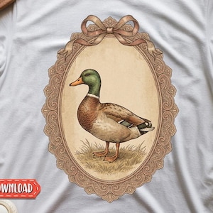 Könnte beinhalten: Ein weißes T-Shirt mit einer zentralen Grafik einer Stockente, die im Gras steht und von einem verzierten, ovalen beigefarbenen Rahmen mit einer dekorativen Schleife umgeben ist. Die Ente hat einen grünen Kopf, einen gelben Schnabel und braun-graues Gefieder.