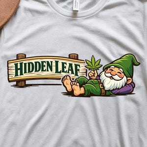 May include: White t-shirt featuring a cartoon gnome holding a cannabis leaf, resting near a wooden sign that reads "Hidden Leaf." The gnome is dressed in green and purple, with a white beard. The design is a digital download.