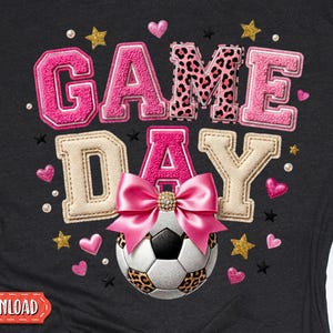May include: Black t-shirt with a pink and leopard print "GAME DAY" design. A soccer ball with a pink bow is featured below the text. Gold stars and pink hearts are scattered around the design. The words "INSTANT DOWNLOAD" and "DIGITAL DOWNLOADS" are also visible.