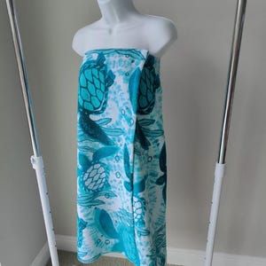 May include: A white beach towel with a turquoise sea turtle pattern. The towel is folded and hanging on a white rack.