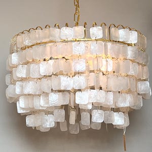 Peut inclure: Un lustre rond à plusieurs niveaux, avec une structure et une chaîne dorées. Il est orné de nombreux éléments carrés, blanc cassé, ressemblant à des coquillages, créant un effet texturé et superposé. Le luminaire est suspendu devant un mur neutre.