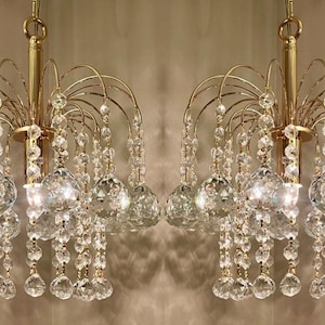 May include: Two gold-toned chandeliers with crystal accents. Each chandelier features a central column with curved arms supporting multiple crystal spheres and dangling crystal strands. The design is symmetrical and elegant.