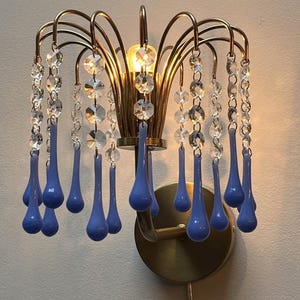 May include: A vintage wall sconce with a brass finish, featuring clear crystal beads and blue teardrop-shaped glass pendants. The light fixture has a decorative, floral-inspired design, adding a touch of elegance to any space.