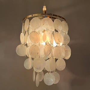 Può includere: Un lampadario decorativo con una montatura in metallo color oro e numerosi dischi rotondi, biancastri, a forma di conchiglia appesi. L'apparecchio è sospeso al soffitto ed emette un bagliore caldo.