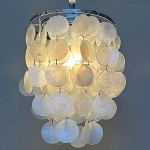 May include: A decorative chandelier with a chrome finish and a cascade of round, off-white shell discs. The light fixture hangs from a single wire and illuminates the room with a warm glow. The chandelier is a unique home decor item.