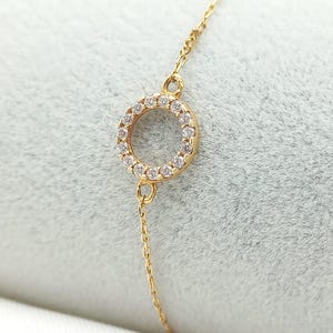 Puede incluir: Un brazalete de cadena dorada con un pequeño colgante circular incrustado con piedras de circonita cúbica transparentes.