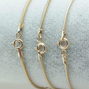 Op de afbeelding: Drie ronde slangenkettingen van goud, elk voorzien van een ronde veerringsluiting. De metalen schakels hebben een gladde, doorlopende textuur. De tekst 14K Round Snake Chain staat onderaan de afbeelding en geeft de materiaalsamenstelling aan.