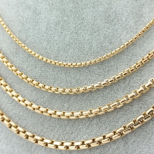 Può includere: Quattro catene a scatola color oro disposte in un motivo semicircolare a cascata. Ogni catena presenta una serie di maglie quadrate interconnesse con una finitura metallica lucida, che mostrano spessori variabili dalla catena superiore a quella inferiore.