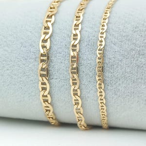 May include: Three gold chains of different lengths and widths, all with a mariner link design. The chains are lying on a gray, textured surface.